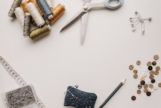 Matériel de couture avec bobines de fil, ciseaux, épingles, boutons et un porte-aiguilles sur une table blanche.