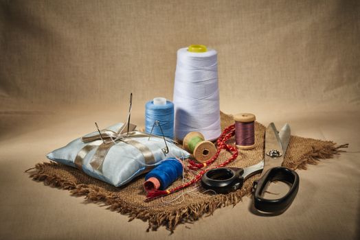 Matériel de couture avec fils colorés, aiguilles, ciseaux et épingles sur un tissu beige. Accessoires faits main.